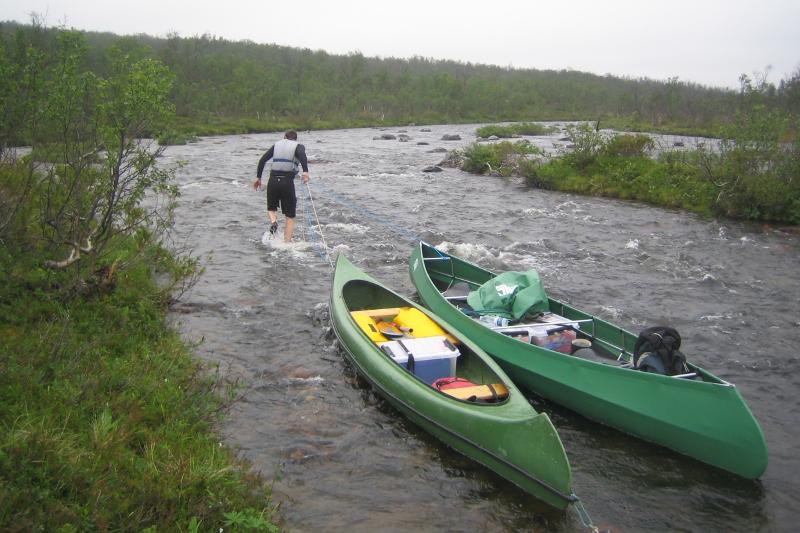 Ein mann dreg to kanoar etter seg opp ei elv. Vått og kaldt vêr.